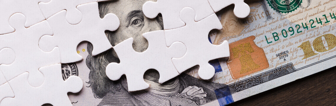 White puzzle pieces on top of a $100 bill, Benjamin Franklin peeking out.
