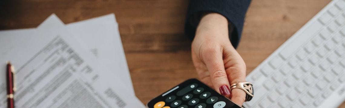 Hand holding a calculator over a desk on which there is paperwork and a computer keyboard