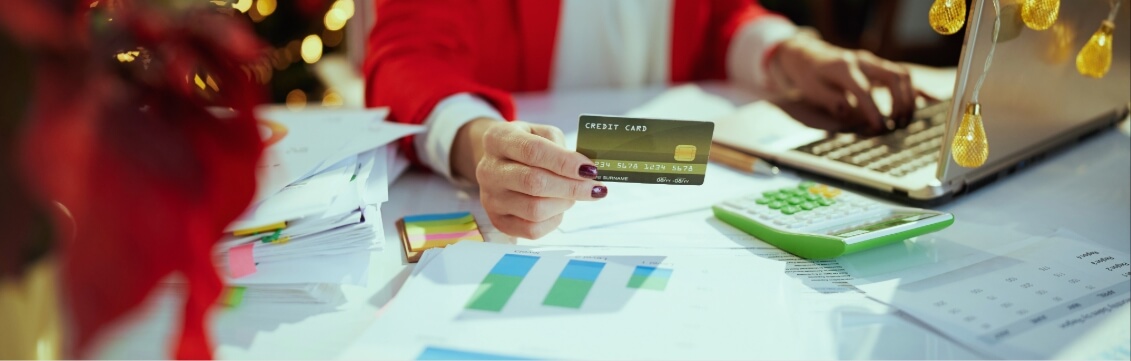 Businesswoman making an online purchase from a laptop, holding a credit card, with holiday lights in the background