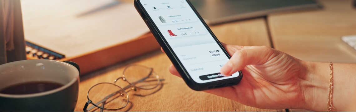 Person completing a mobile checkout for retail items using an ecommerce app, with glasses and coffee on a desk nearby